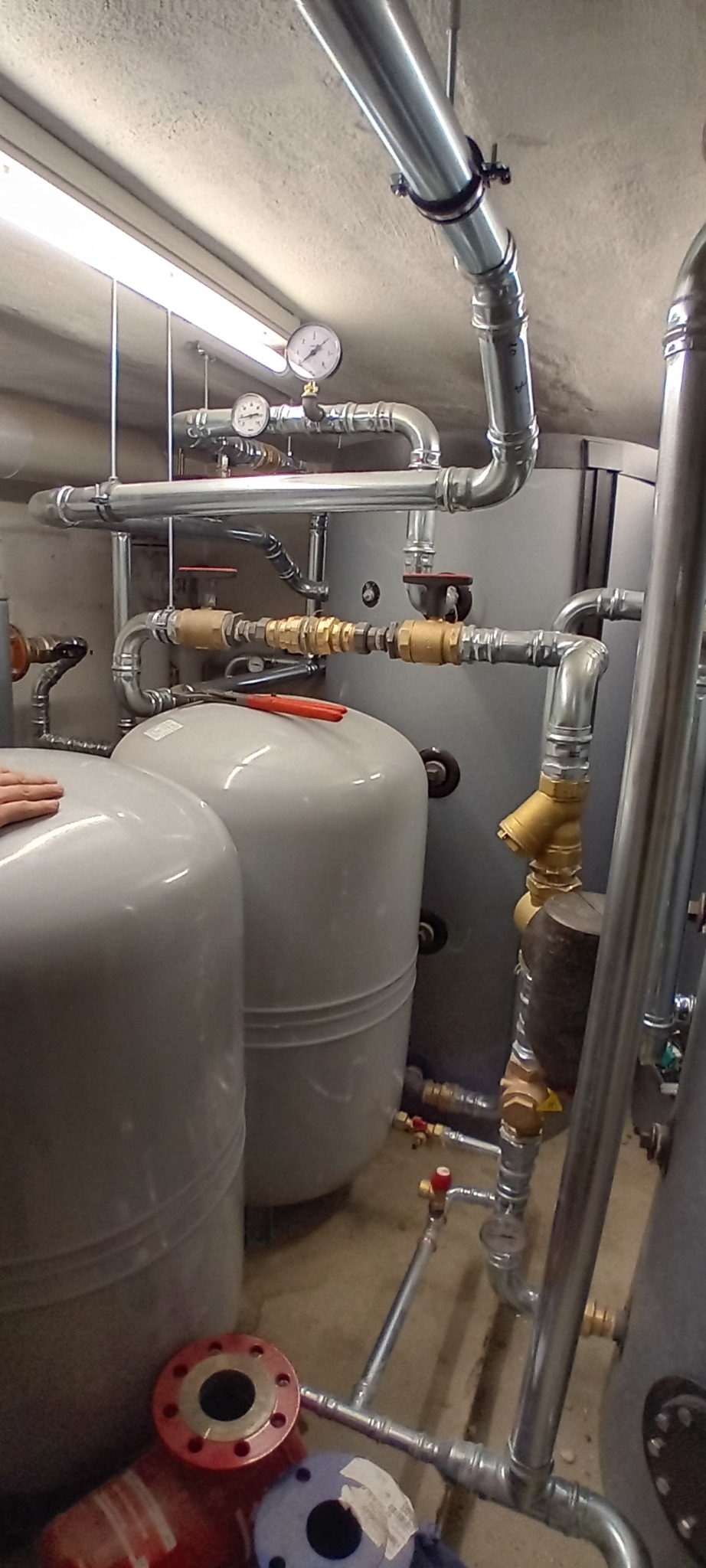 Szare zbiorniki ciśnieniowe i rury z zaworami w kotłowni. Na rurach widoczne manometry. Na zbiornikach leżą czerwone kombinerki. Na podłodze kołnierze.