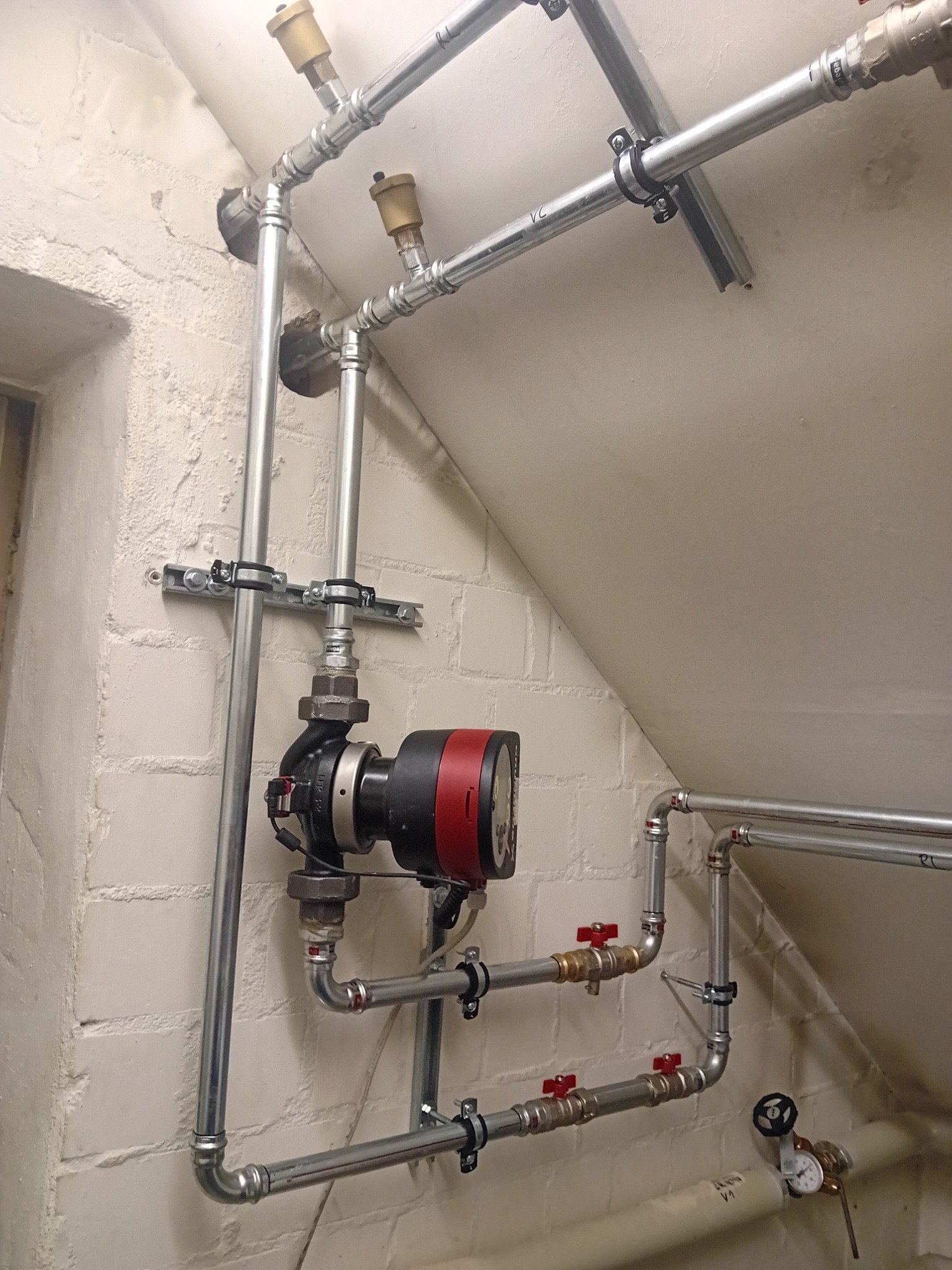 Szare rury instalacji CO z czerwonymi zaworami i pompą obiegową na tle białej ściany z cegły i skośnego sufitu. Widoczne mocowania do ściany i sufitu.