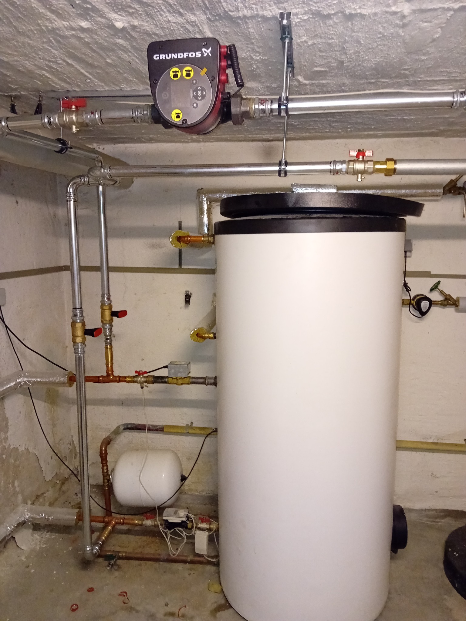 Biały bojler z czarną pokrywą i pompą obiegową Grundfos, zamontowany w piwnicy z widocznymi rurami i zaworami. Instalacja grzewcza w pomieszczeniu technicznym.