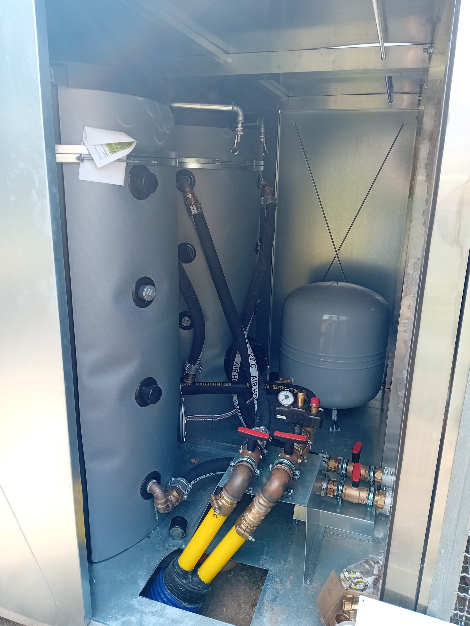 Wnętrze metalowej szafy z zamontowanym szarym zbiornikiem wyrównawczym, rurami miedzianymi i czarnymi przewodami hydraulicznymi z żółtymi osłonami.