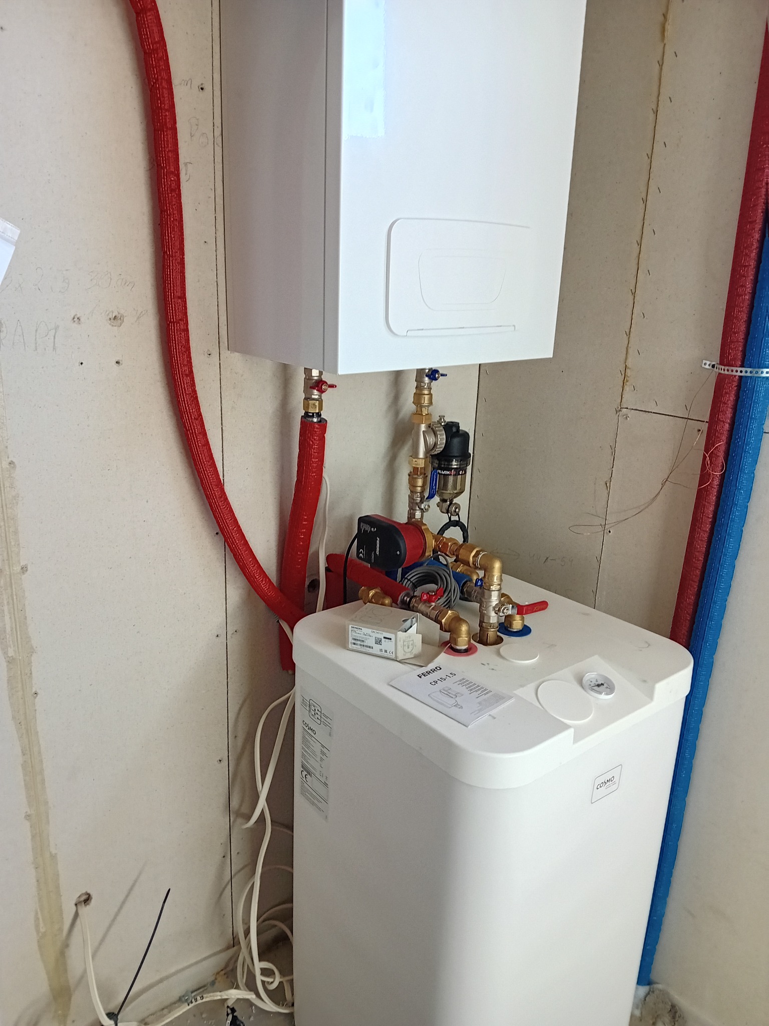 Biały piec CO Cosmo z czerwonymi rurami w izolacji, zamontowany na ścianie z widocznymi śladami po instalacji. Widoczne elementy armatury i pompy obiegowej.