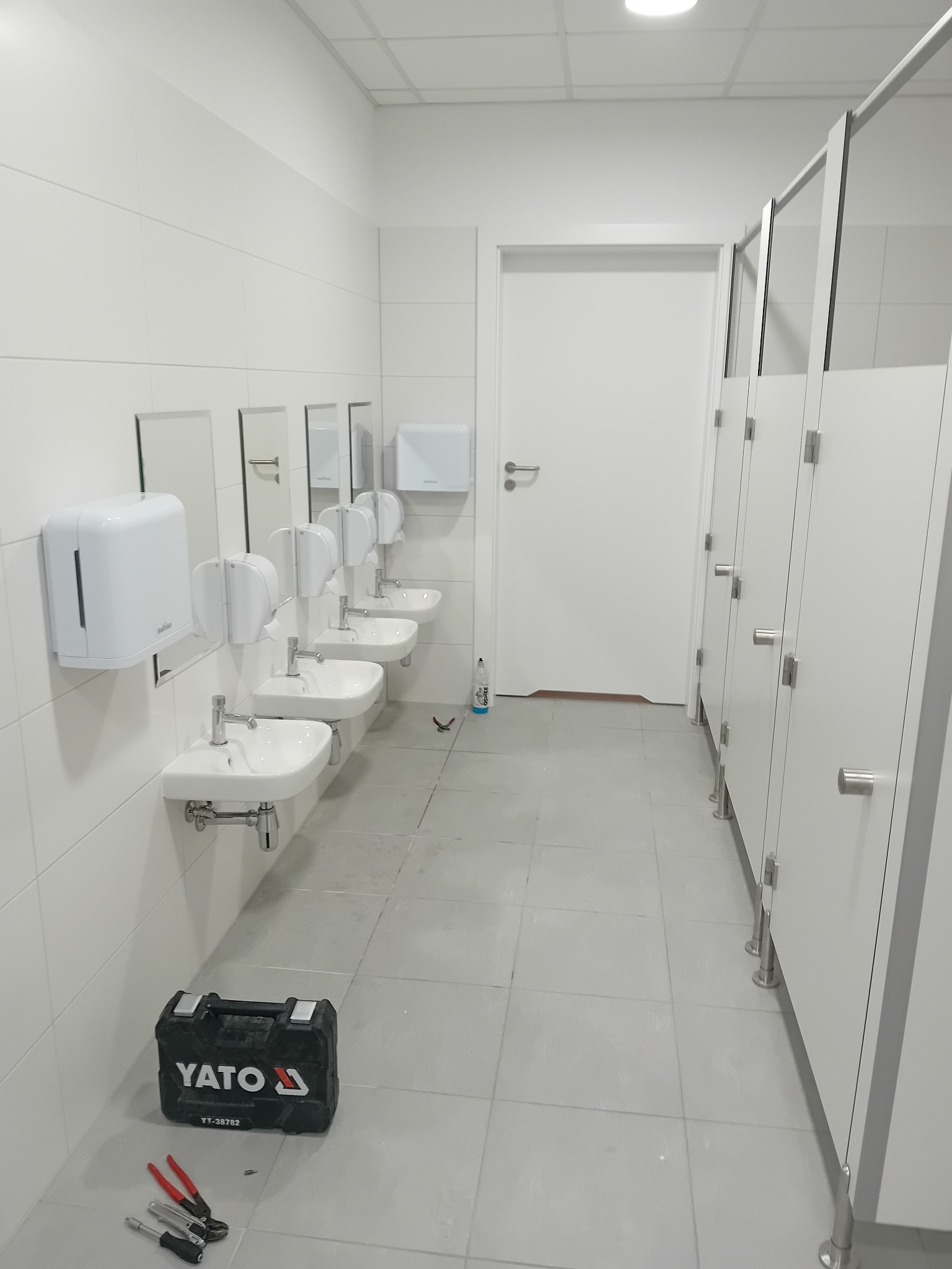 Biała łazienka publiczna po montażu armatury w Starogardzie Gdańskim. Widoczne umywalki, dozowniki mydła i kabiny WC. Na podłodze leży skrzynka narzędziowa.