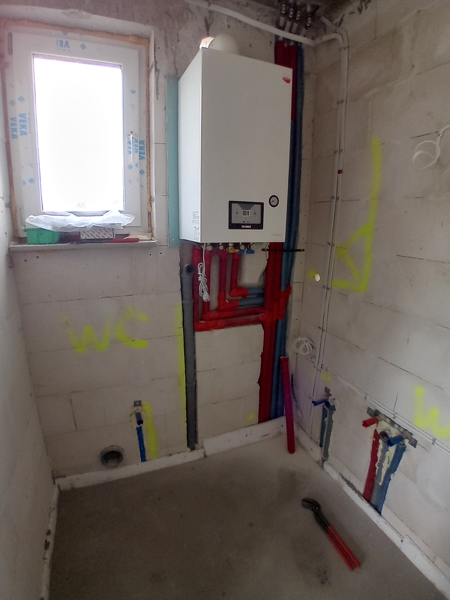 Biały piec gazowy Termet zamontowany na ścianie z instalacją hydrauliczną w surowym pomieszczeniu. Widoczne rury w kolorach czerwonym i niebieskim oraz narzędzie hydrauliczne.