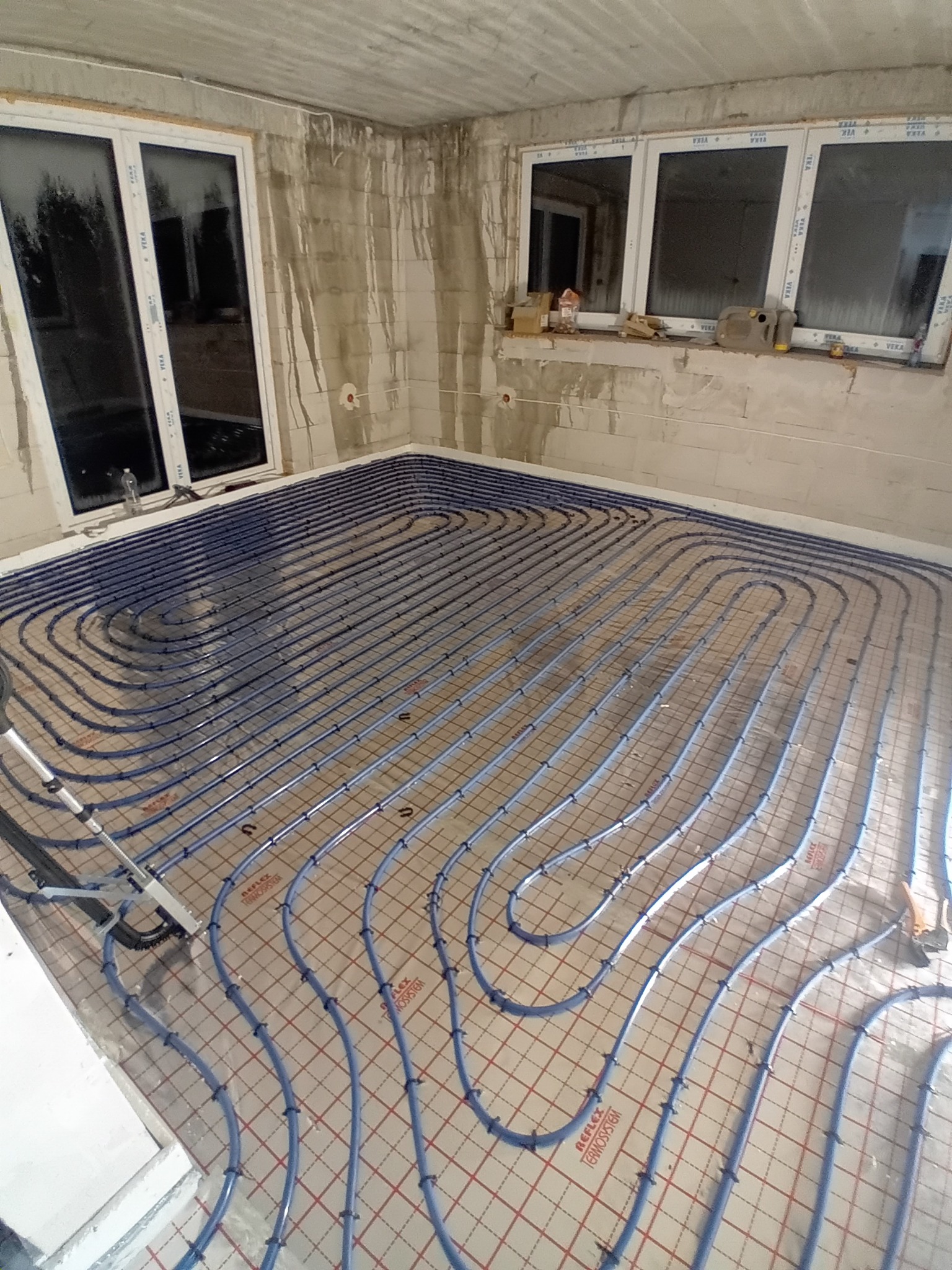 Niebieskie rury ogrzewania podłogowego ułożone na siatce w surowym wnętrzu z dwoma oknami. Widoczne narzędzia i materiały budowlane. Etap instalacji przed wylewką.