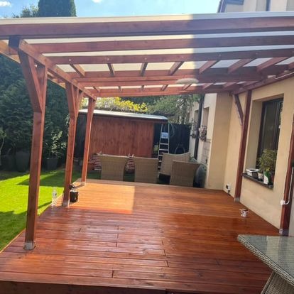 Zadaszony taras z drewnianą podłogą i konstrukcją, widok na ogród z elementami mebli ogrodowych. Świeżo polakierowane deski podłogowe.