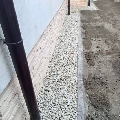 Drenaż opaskowy z jasnych kamieni przy ścianie budynku z brązową rurą spustową i kostką brukową. Widok z góry, lekki kąt.