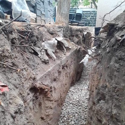 Wykop z drenażem żwirowym w ziemi, widok z góry. Fragment drzewa i budynku w tle. Prace ziemne przy instalacji sanitarnej.