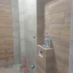 Henryk Olesiński - Wnętrze łazienki w trakcie prac wykończeniowych. Widoczne płytki imitujące drewno na ścianach, częściowo zamontowana toaleta i narzędzia glazurnicze.