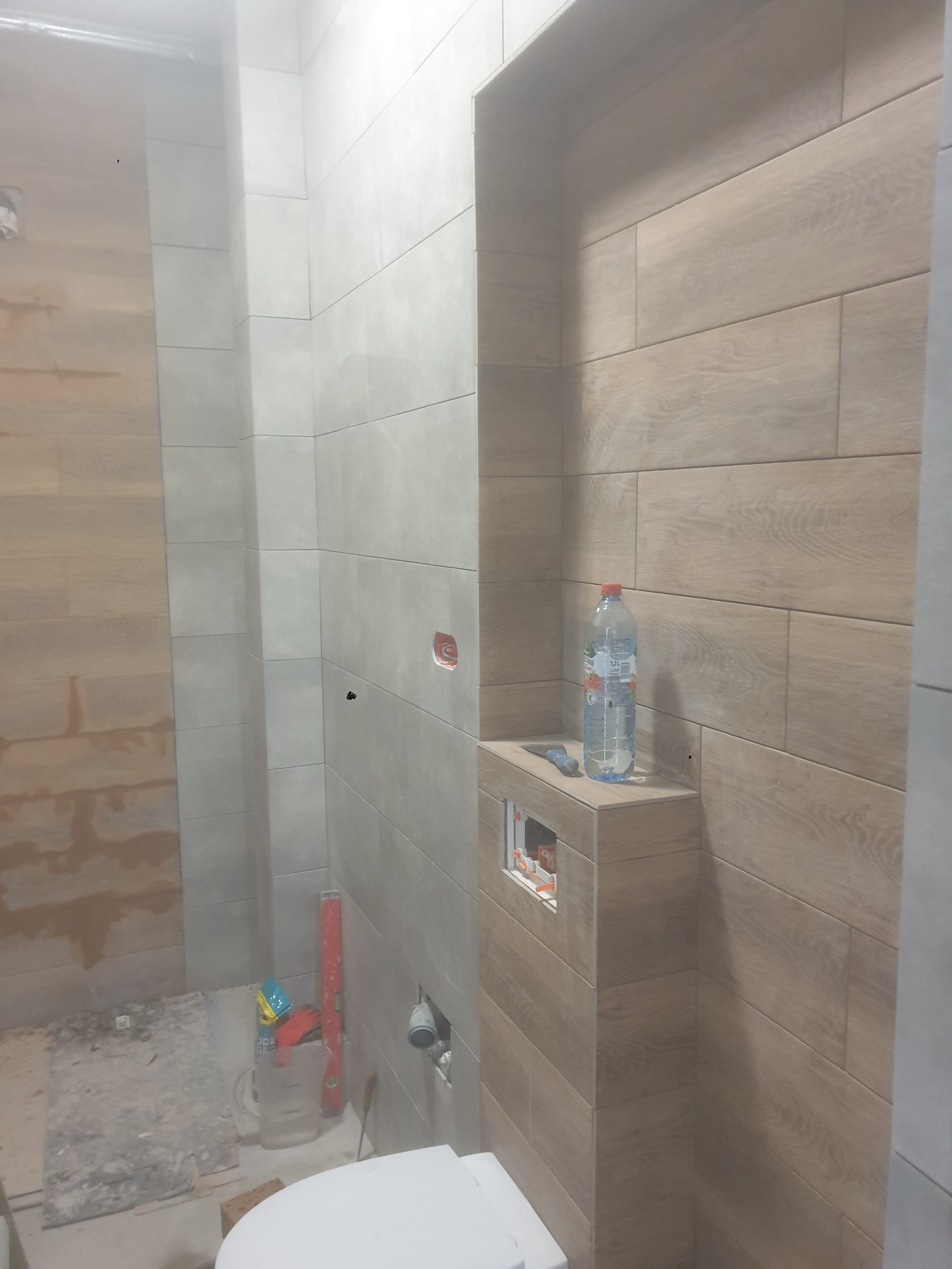 Wnętrze łazienki w trakcie prac wykończeniowych. Widoczne płytki imitujące drewno na ścianach, częściowo zamontowana toaleta i narzędzia glazurnicze.