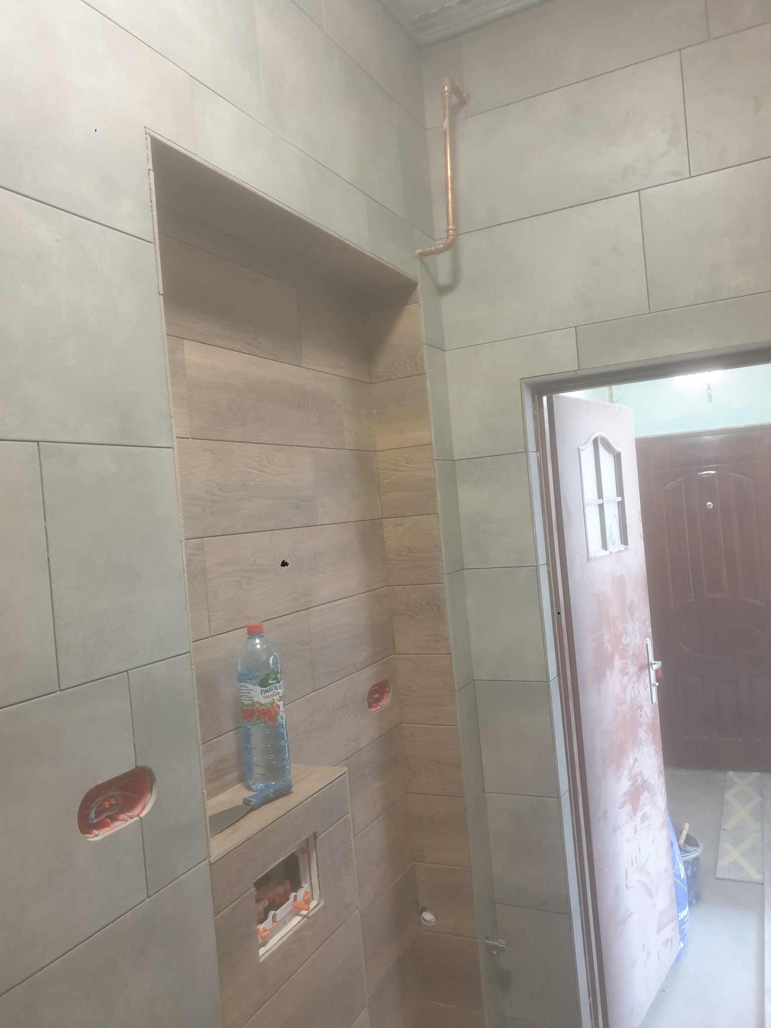 Ściana w łazience wyłożona płytkami imitującymi drewno, z wnęką, w której stoi butelka wody. Widoczne otwory na instalacje i fragment rury miedzianej.