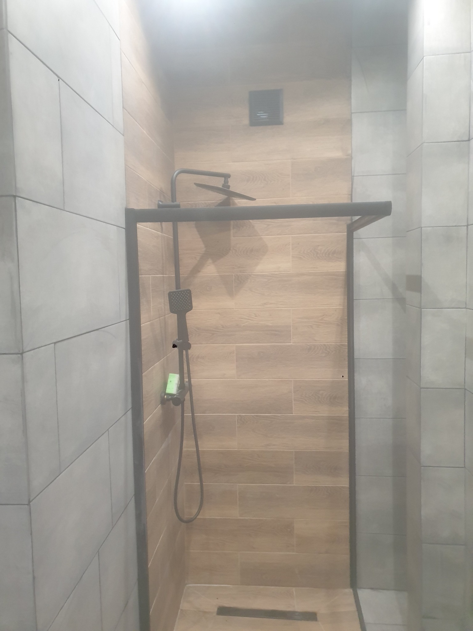 Nowoczesna kabina prysznicowa z czarną armaturą i płytkami imitującymi drewno. Minimalistyczny design, szklana ściana, widoczny odpływ liniowy.