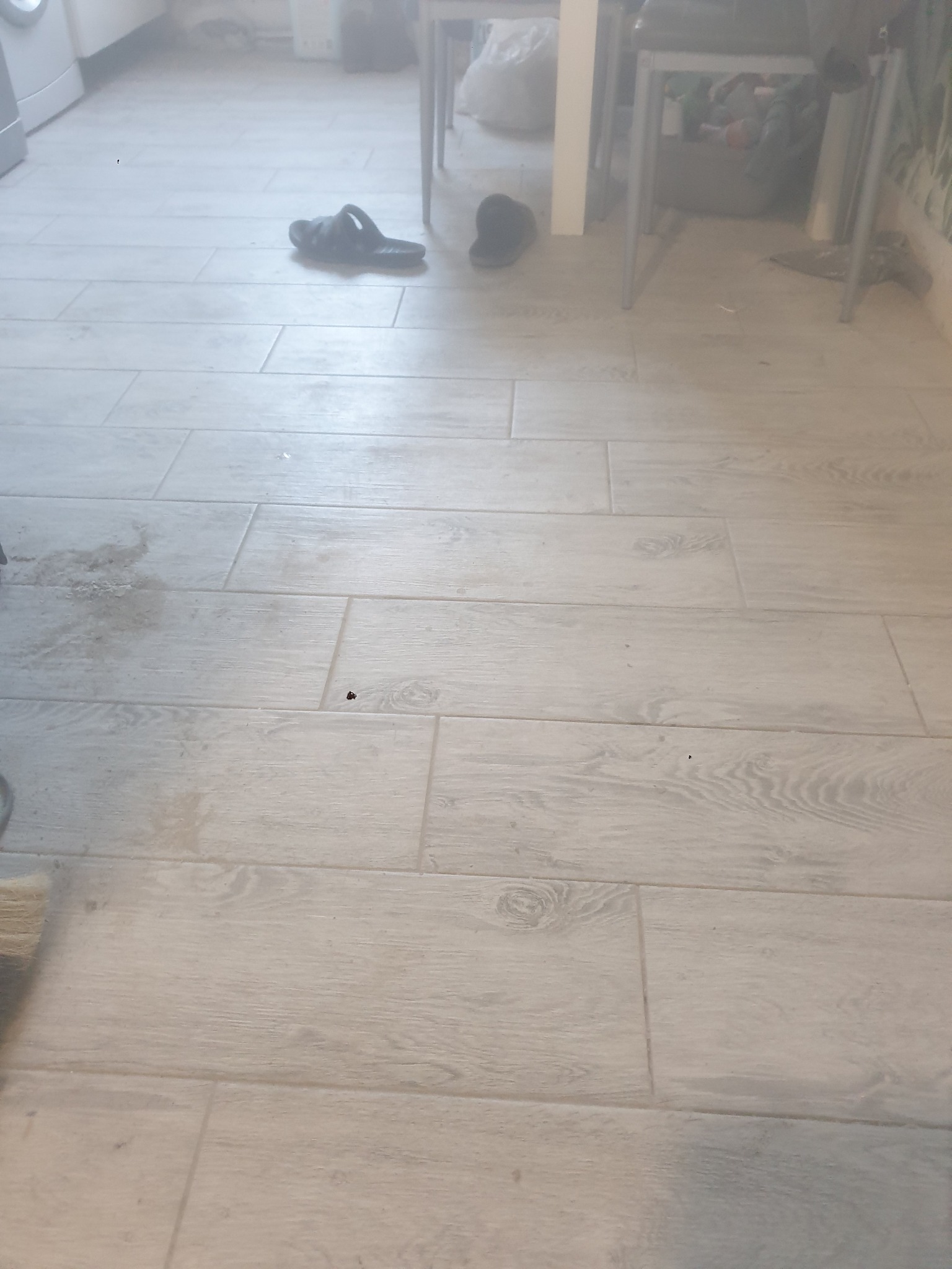 Jasne, drewnopodobne panele podłogowe ułożone w kuchni. W tle widoczne krzesła i przedmioty codziennego użytku. Minimalistyczny styl wykończenia.