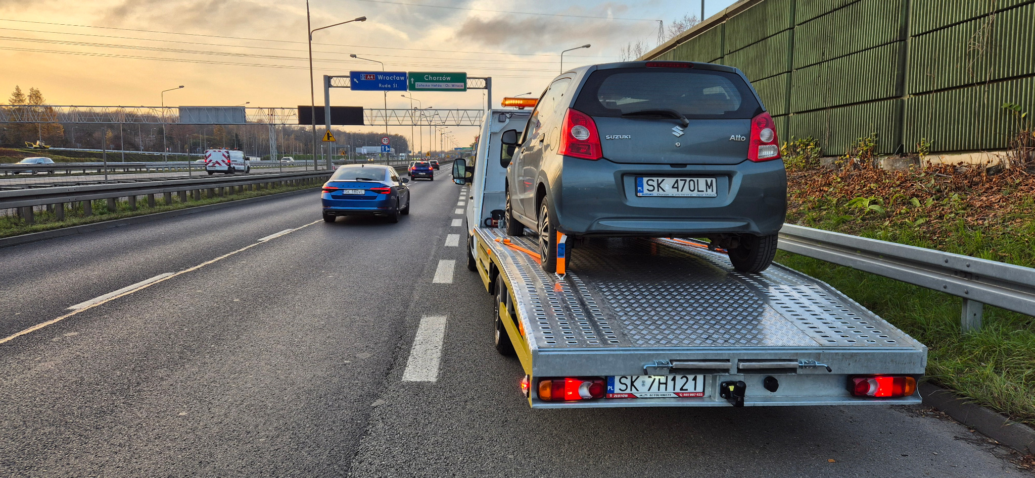 Szary samochód Suzuki Alto zabezpieczony pasami na platformie pomocy drogowej. Widok z tyłu, tablice rejestracyjne widoczne. Autostrada A4 w tle.