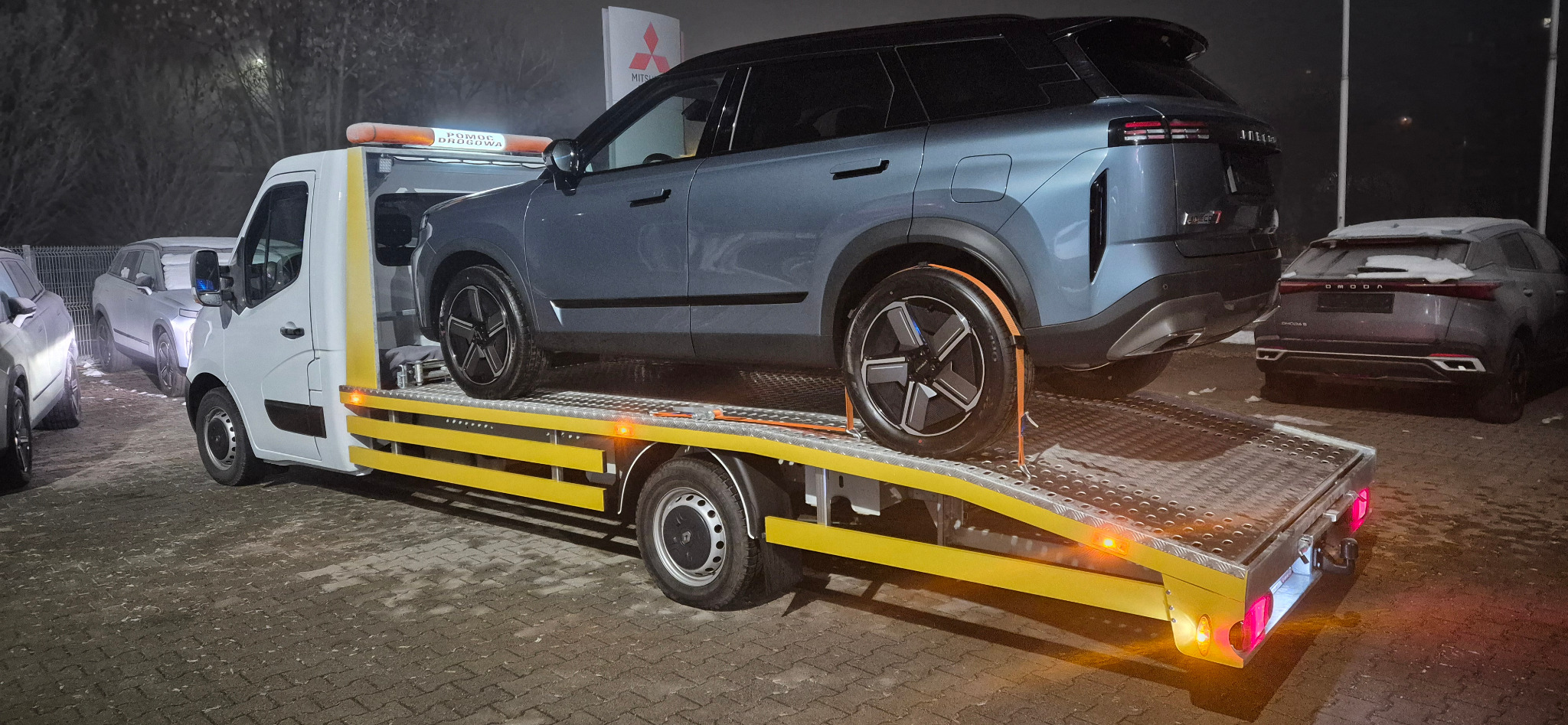 Niebieski SUV Omoda na żółtej lawecie pomocy drogowej, widok z tyłu. W tle salon samochodowy Mitsubishi i inne pojazdy Omoda. Nocne oświetlenie.