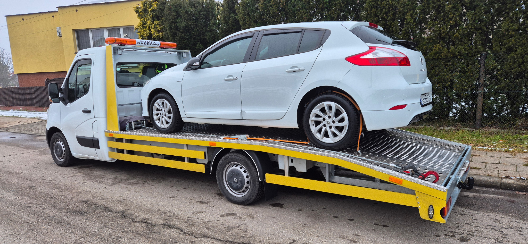 Biały samochód osobowy, Renault Megane, zabezpieczony pasami transportowymi na platformie lawety pomocy drogowej z żółtymi elementami. Widok z boku, częściowo ośnieżone otoczenie.