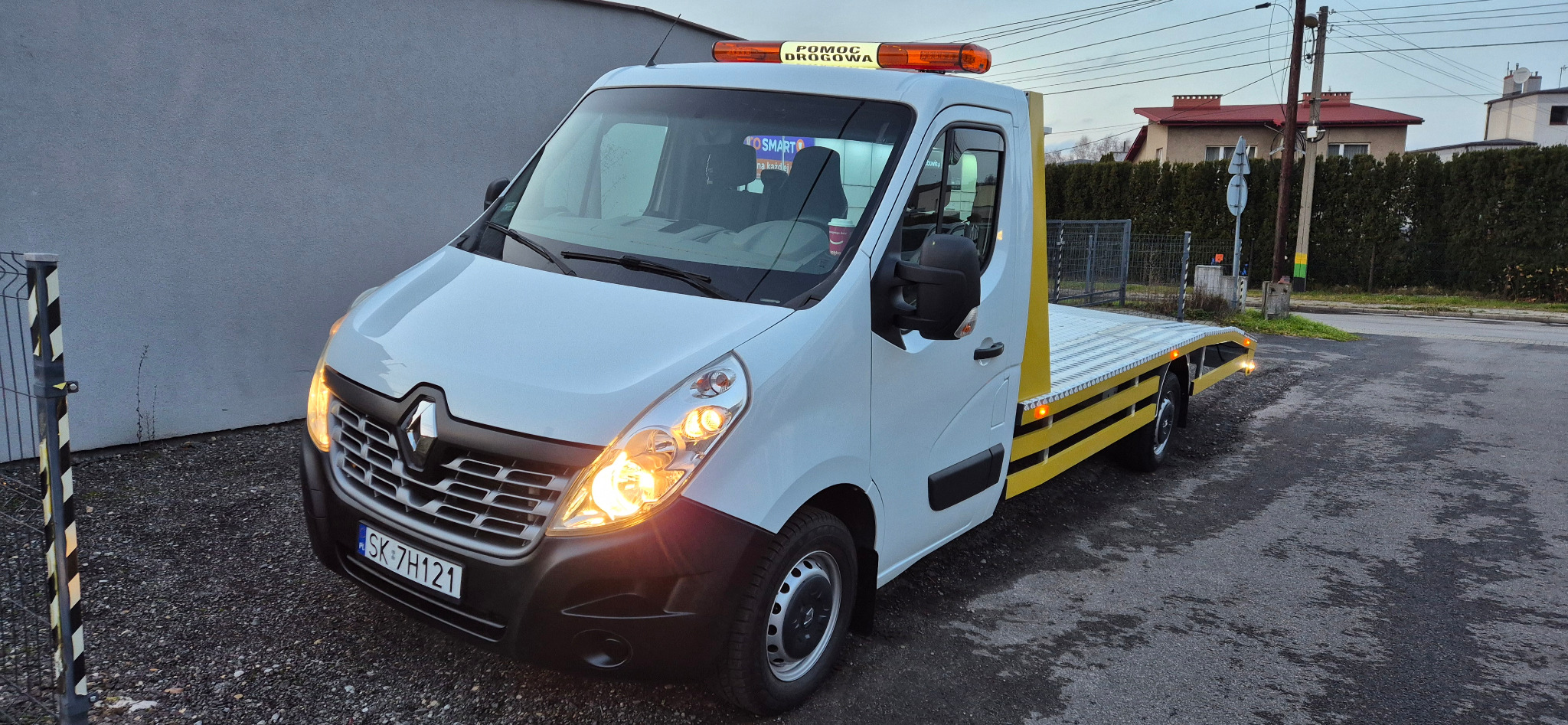Biała laweta Renault Master z żółtymi elementami, zaparkowana na ulicy. Włączone światła mijania i pomarańczowe światło ostrzegawcze na dachu.