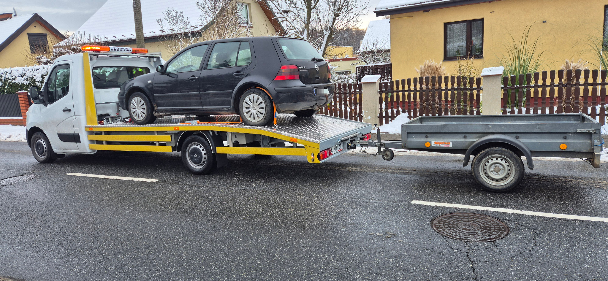 Czarny samochód osobowy na żółtej platformie autolawety z przyczepą na tle zimowego krajobrazu z domami. Widoczny napis 'Pomoc Drogowa' na dachu pojazdu.