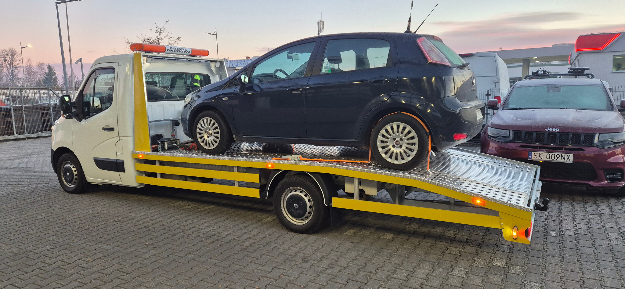 Czarny samochód osobowy zabezpieczony pasami na żółtej platformie lawety pomocy drogowej, na parkingu, w tle zaparkowany czerwony Jeep i budynek stacji benzynowej.