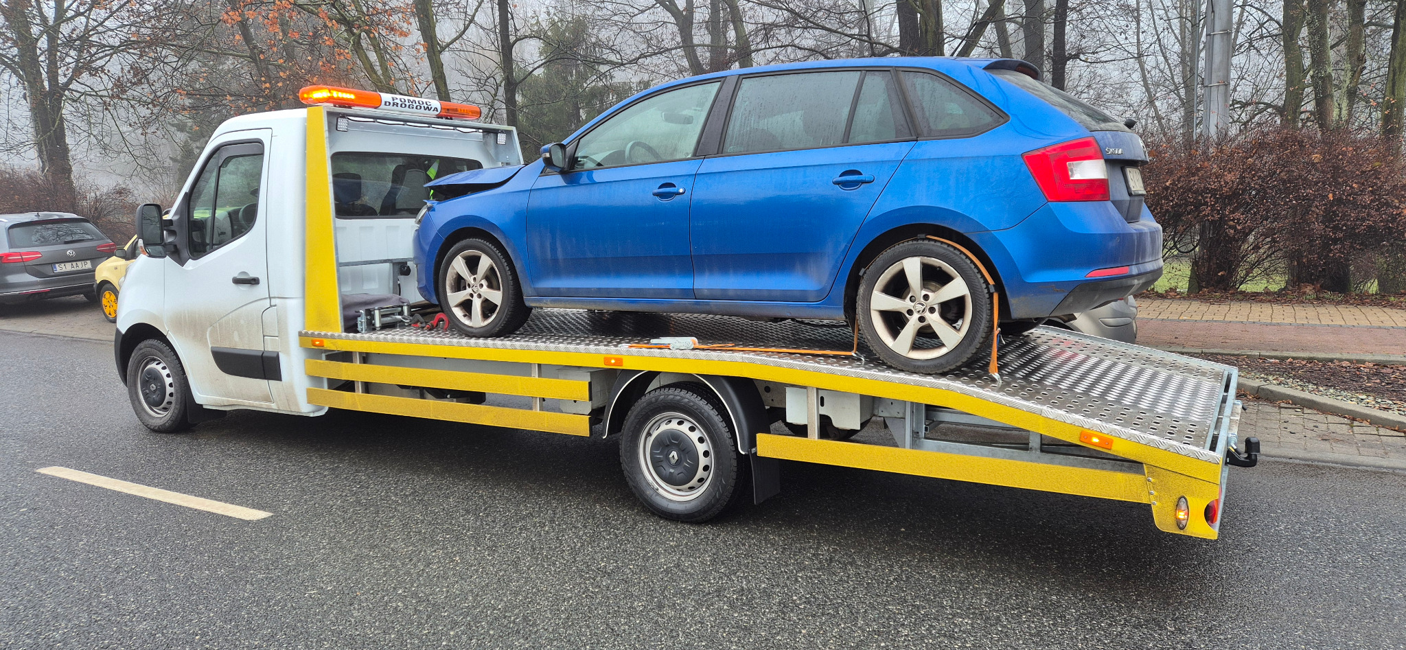 Niebieski samochód z uszkodzonym przodem, zabezpieczony pasami, transportowany na żółtej lawecie pomocy drogowej na tle jesiennego krajobrazu.