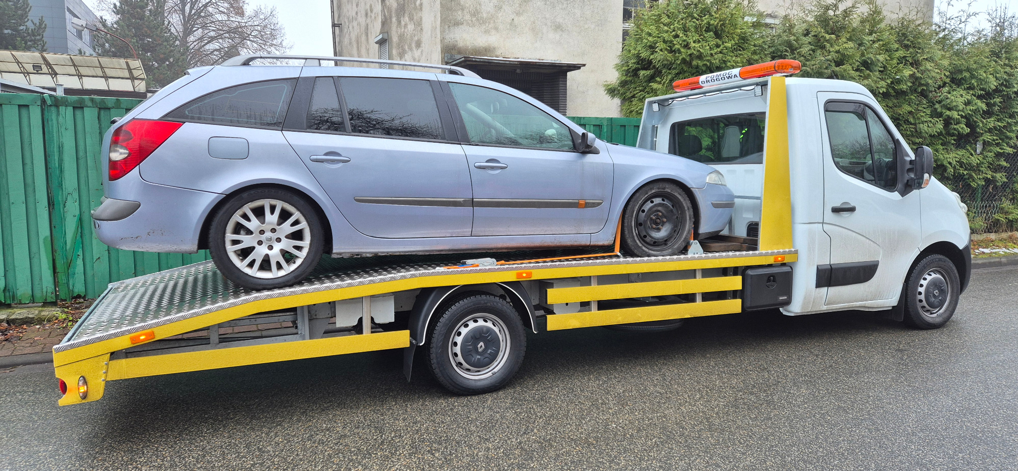 Niebieskie kombi na żółtej platformie lawety, jedno koło uszkodzone. Widoczny budynek w tle i zielony żywopłot. Pomoc drogowa w trakcie transportu pojazdu.