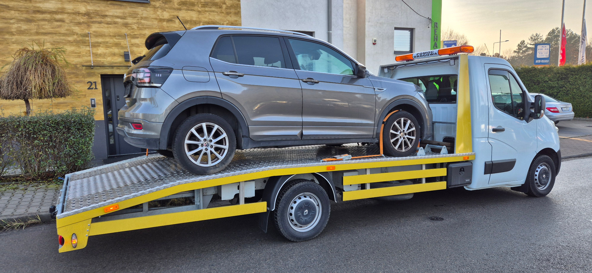 Szary samochód osobowy Volkswagen T-Cross zabezpieczony pasami transportowymi na platformie lawety pomocy drogowej, na tle budynku z numerem 21.