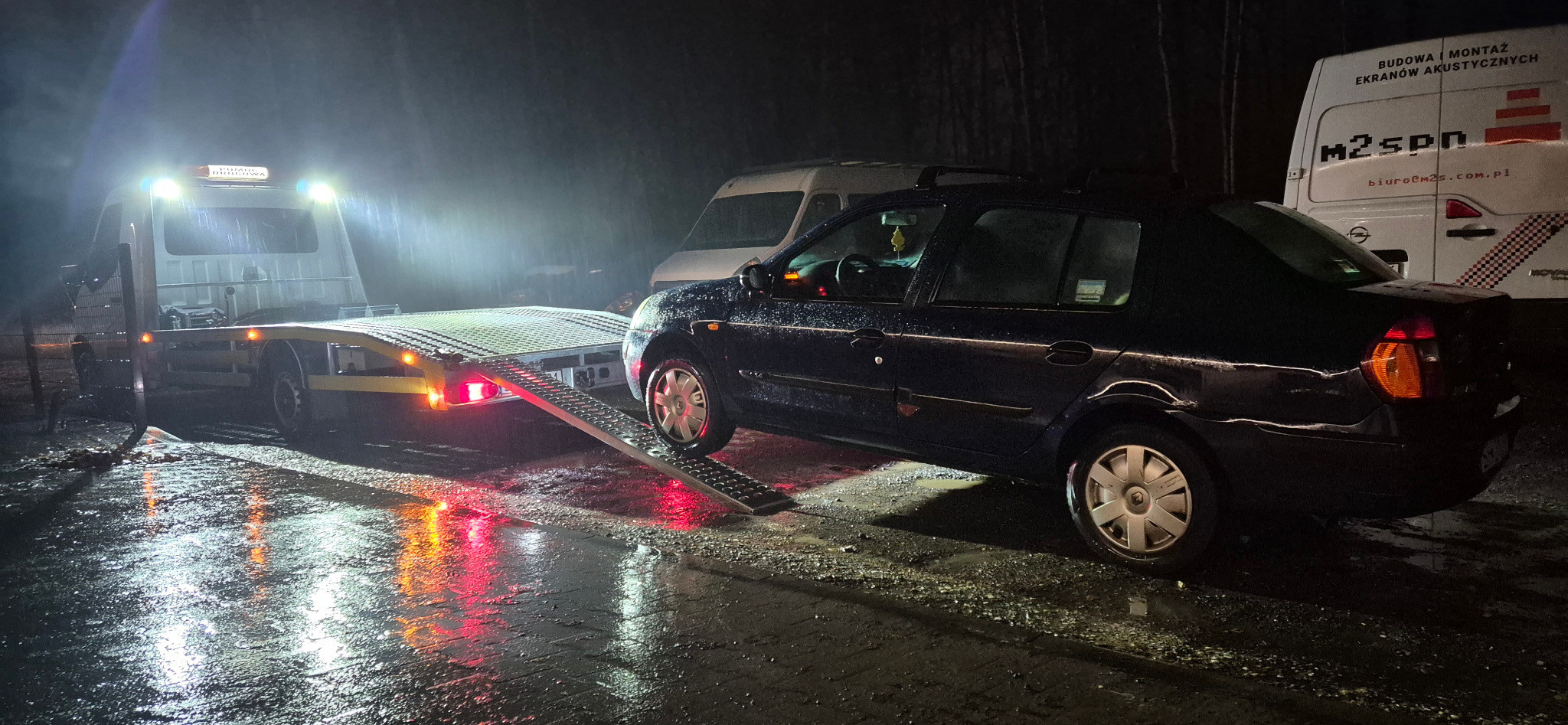 Nocne załadowywanie ciemnego samochodu osobowego na platformę pojazdu pomocy drogowej. Mokra nawierzchnia odbija światła. W tle inne pojazdy.