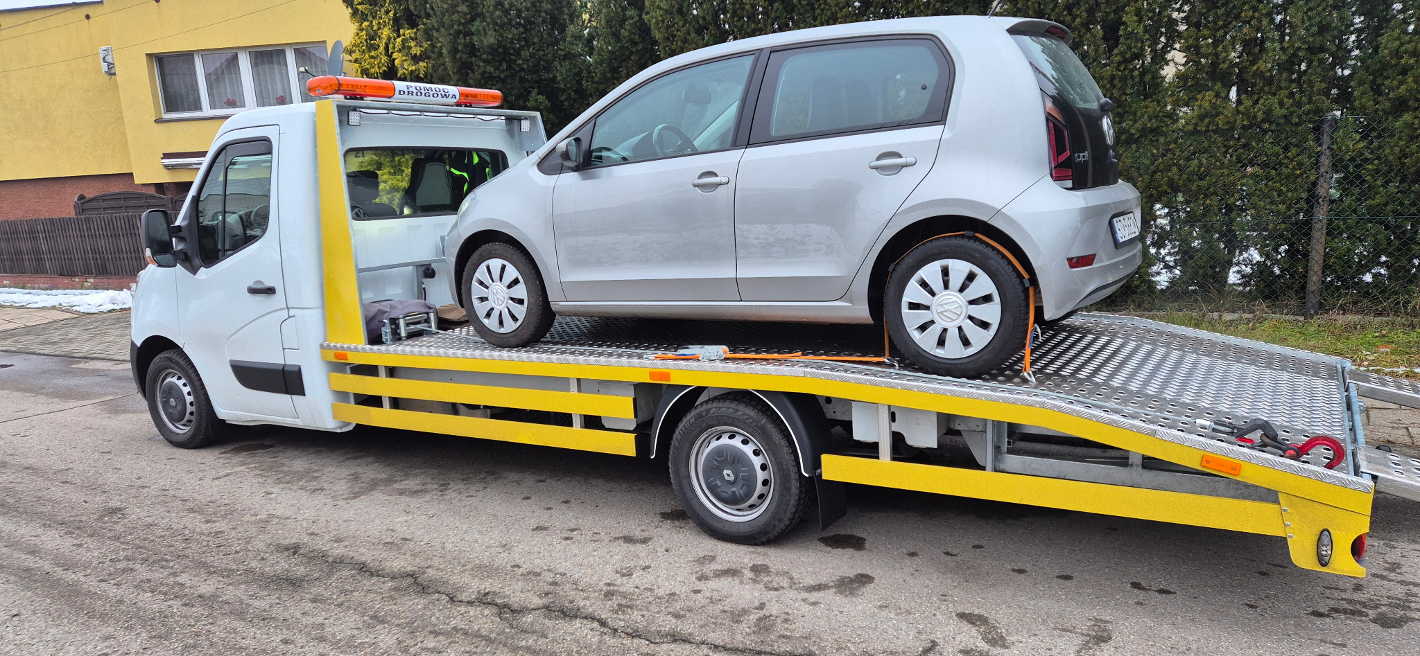 Laweta z srebrnym Volkswagenem Up! zabezpieczonym pasami transportowymi. Widoczny fragment budynku i zielone krzewy w tle. Pomoc drogowa w trakcie akcji.