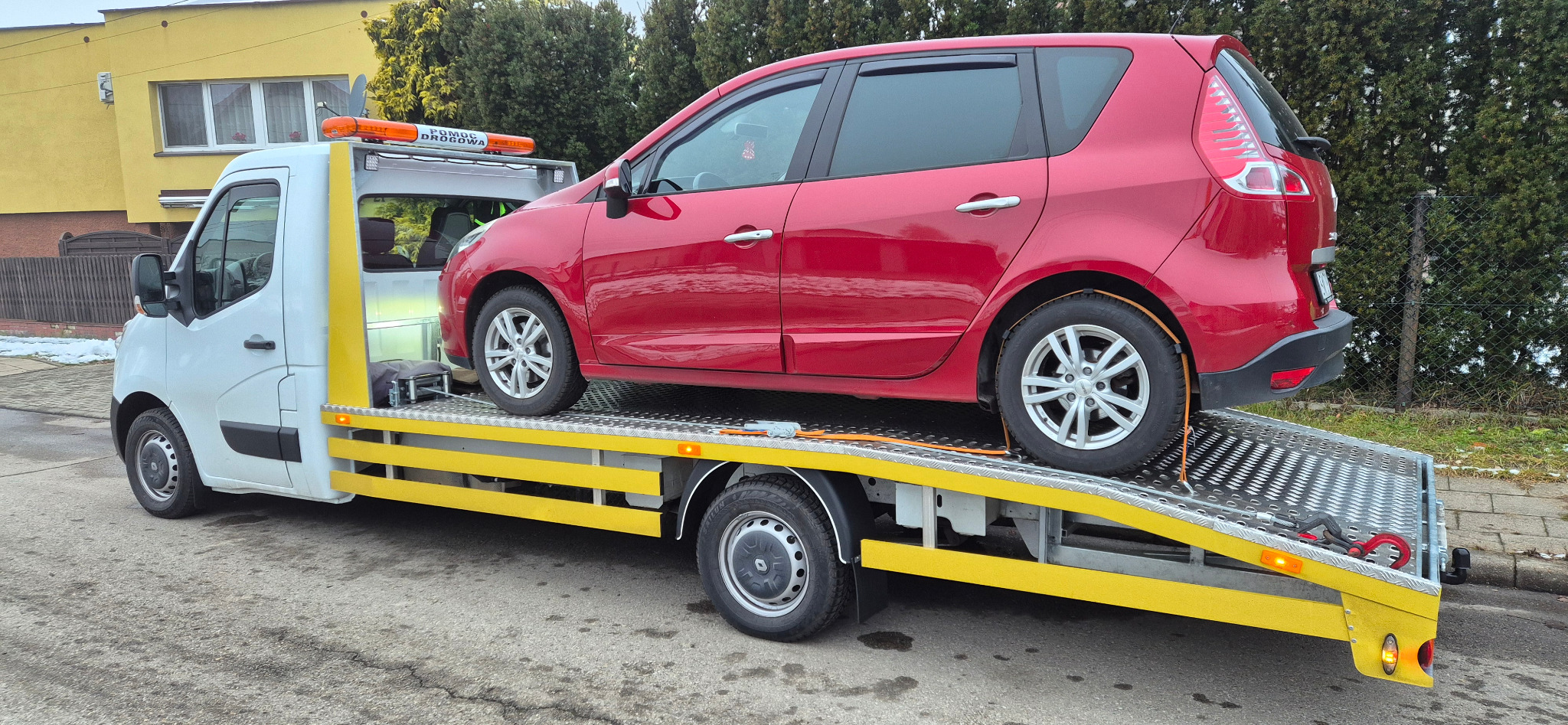 Czerwony samochód osobowy zabezpieczony pasami na platformie białej lawety z żółtymi elementami, widok z boku na tle ulicy i zabudowań.