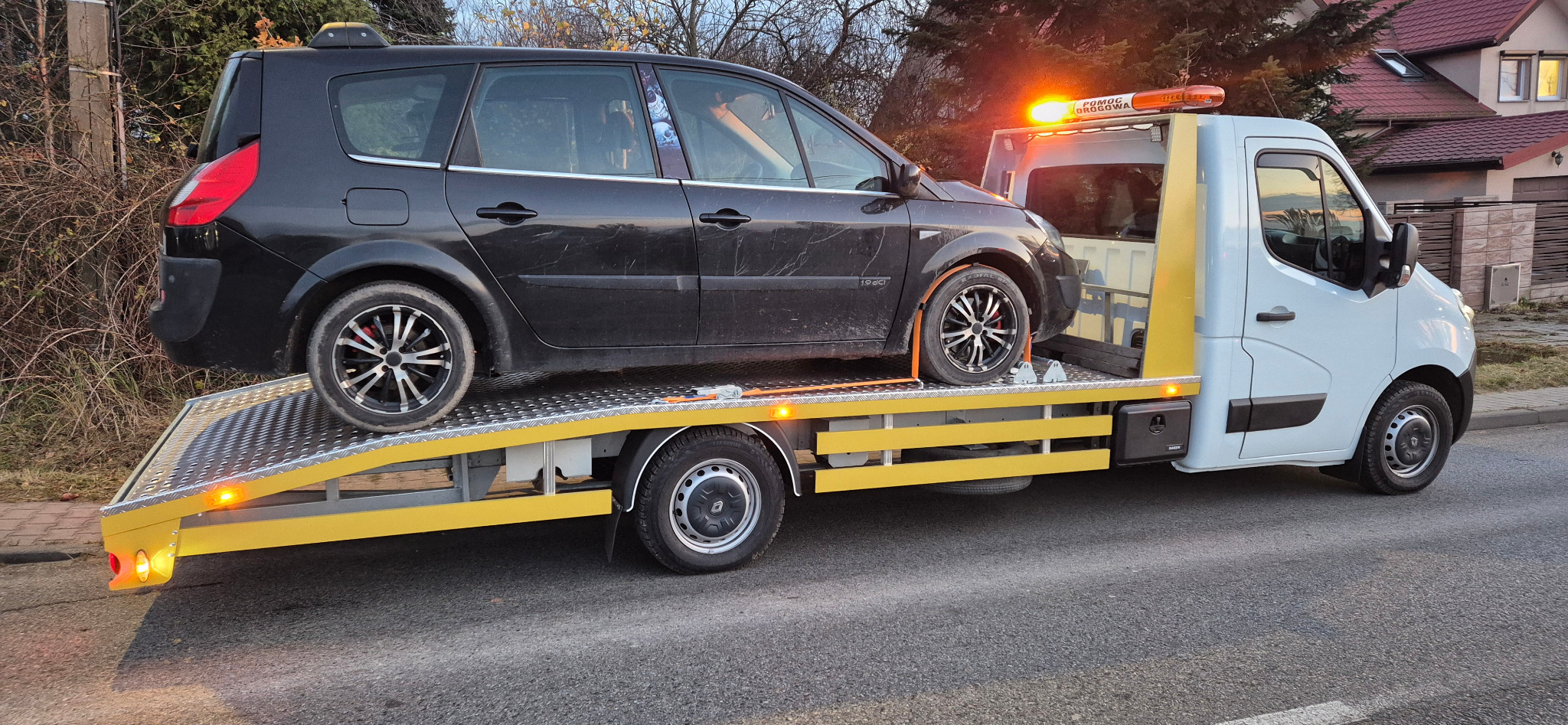 Czarny samochód osobowy z felgami na żółtej platformie lawety, światło ostrzegawcze włączone, widok z boku na tle zabudowań i drzew.