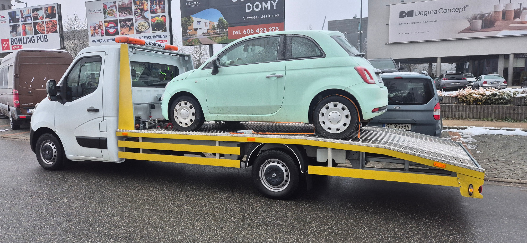 Laweta z jasnozielonym Fiatem 500 na platformie, widok z boku na tle miejskiego krajobrazu z reklamami. Auto zabezpieczone pasami transportowymi.