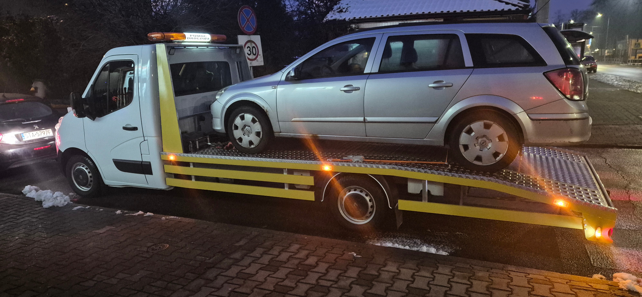 Srebrny samochód osobowy typu kombi na żółtej platformie pojazdu pomocy drogowej z włączonymi światłami ostrzegawczymi na tle miejskiej scenerii.