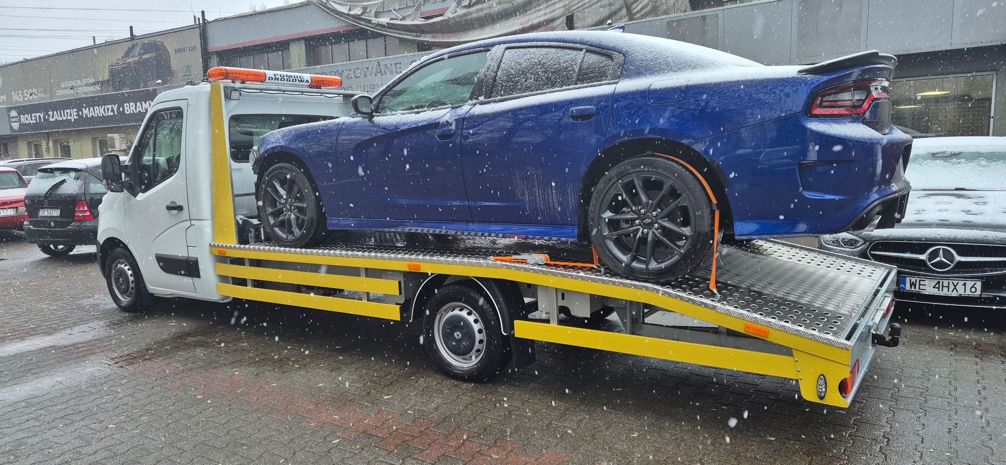 Niebieski samochód Dodge Charger zabezpieczony pasami na żółtej lawecie pomocy drogowej podczas opadów śniegu. Widoczny fragment budynku w tle.
