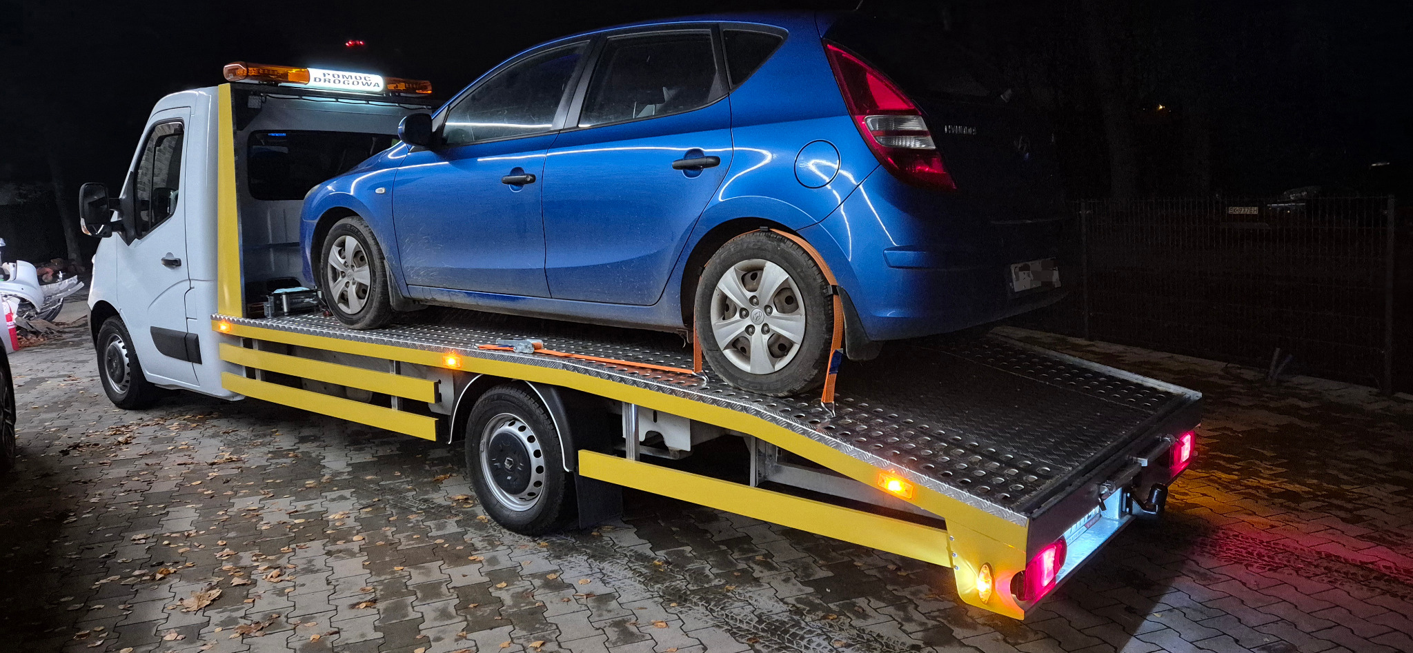 Nocne ujęcie niebieskiego auta zabezpieczonego pasami na platformie lawety z włączonymi światłami ostrzegawczymi, sygnalizującymi pomoc drogową.