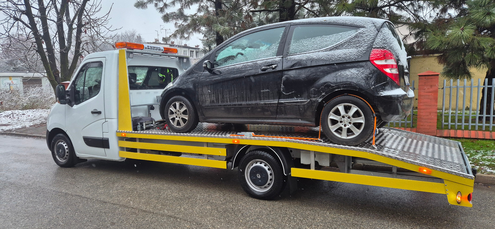 Czarny samochód osobowy zabezpieczony pasami na żółtej platformie lawety pomocy drogowej w zimowy dzień. Widoczny budynek w tle.