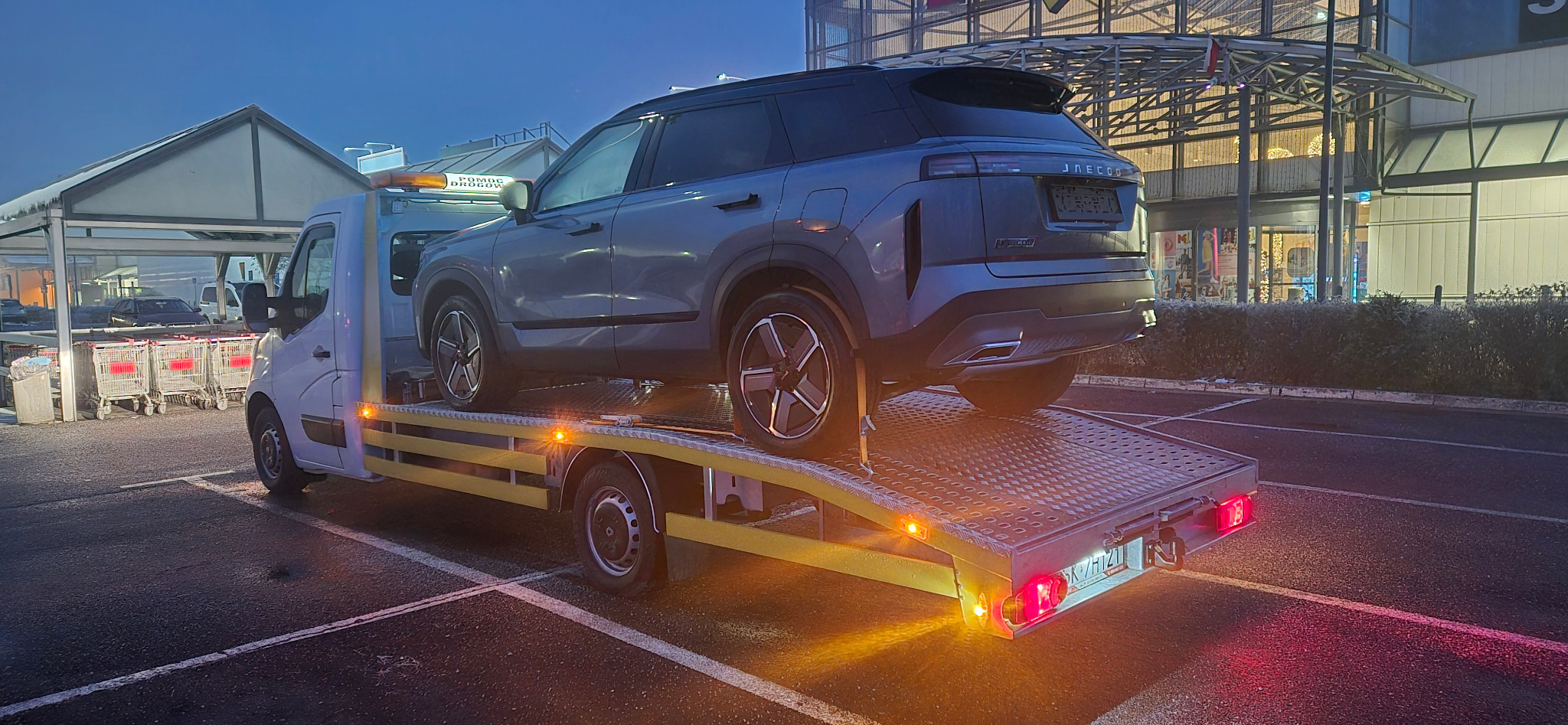 Szary SUV marki JAECOO na platformie białej lawety z żółtymi pasami, na parkingu przed centrum handlowym o zmierzchu. Widoczny napis 'Pomoc Drogowa'.