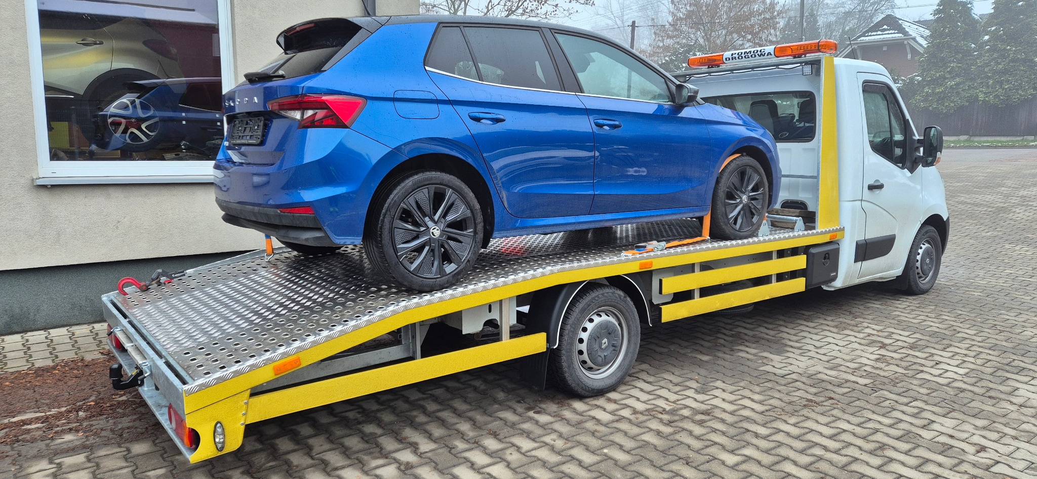 Niebieska Skoda na żółto-srebrnej platformie lawety z napisem 'Pomoc Drogowa' na tle budynku i drzew. Auto zabezpieczone pasami transportowymi.