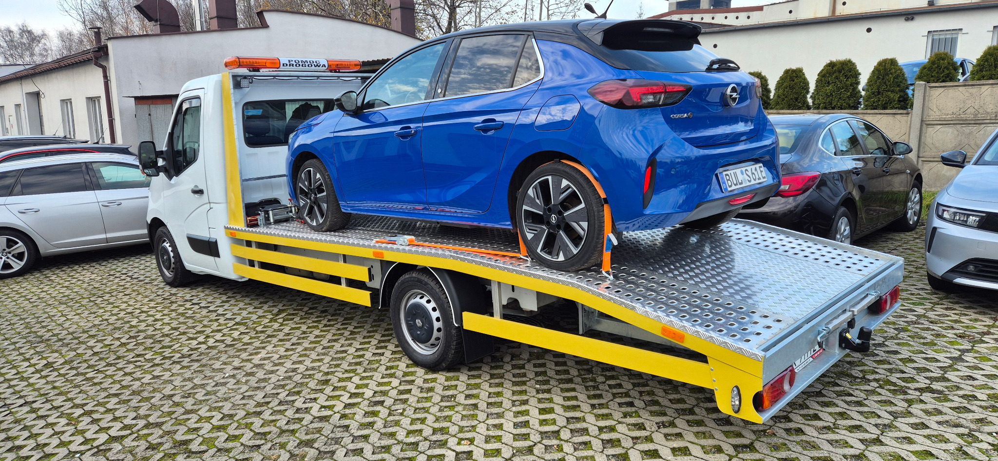 Niebieska Opel Corsa zabezpieczona pasami na platformie białej lawety z żółtymi elementami, zaparkowanej na kostce brukowej. Widoczny budynek w tle.