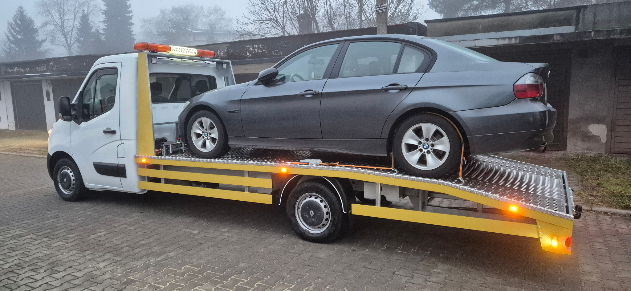 Szara osobówka BMW na żółtej platformie lawety z pomarańczowym światłem ostrzegawczym na tle szarej kostki brukowej i zarośli w pochmurny dzień.