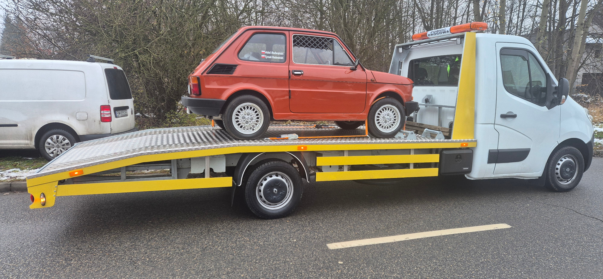 Pomarańczowy Fiat 126p z klatką na lawecie pomocy drogowej. W tle biały bus i drzewa. Widoczny napis 'Pomoc Drogowa' na pojeździe.