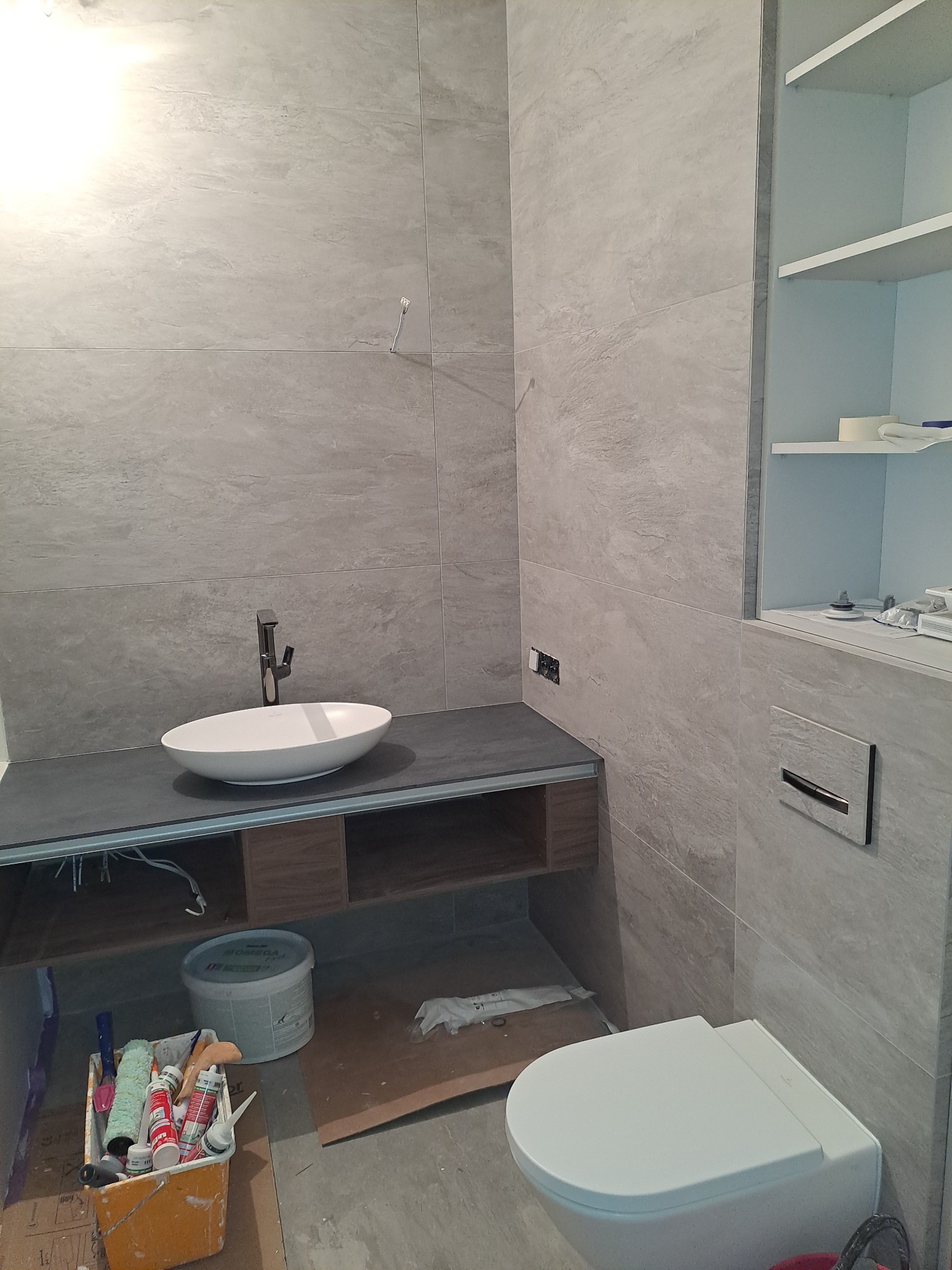 Nowoczesna łazienka w szarościach z umywalką nablatową, toaletą podwieszaną i zabudową. Widoczne narzędzia i materiały sugerują etap prac wykończeniowych.