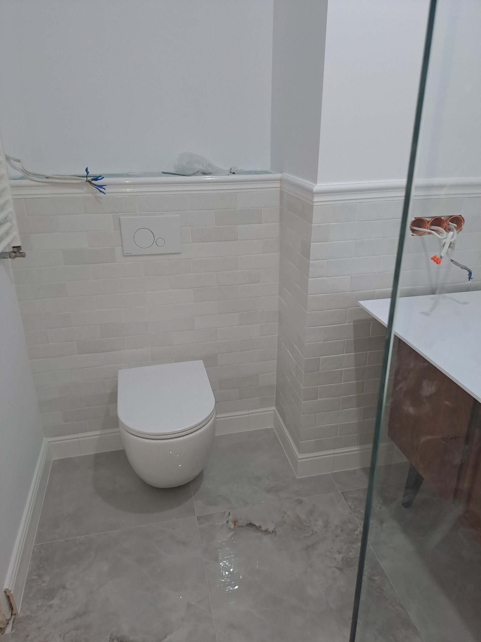 Jasna łazienka po remoncie: białe płytki imitujące cegłę, podwieszana toaleta, szara podłoga z płytek imitujących marmur, częściowo widoczna szklana kabina.