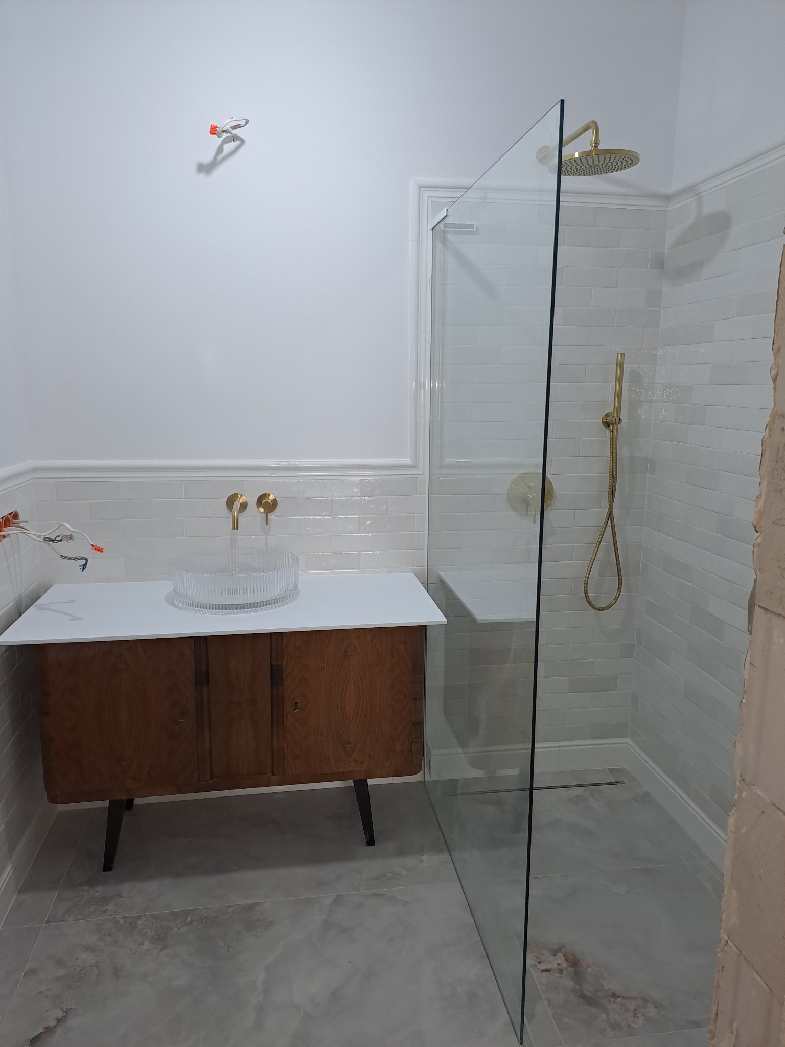 Nowoczesna łazienka z kabiną prysznicową typu walk-in, umywalką na szafce z ciemnego drewna i złotą armaturą. Widoczne niedokończone podłączenia elektryczne.