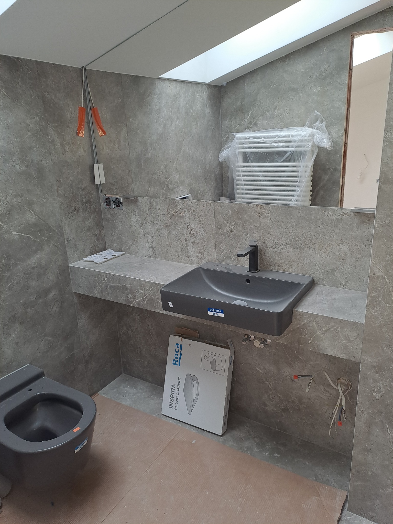 Nowoczesna łazienka w szarościach: umywalka nablatowa, toaleta Roca i grzejnik drabinkowy w folii ochronnej. Widoczne przewody elektryczne i płytki imitujące marmur.