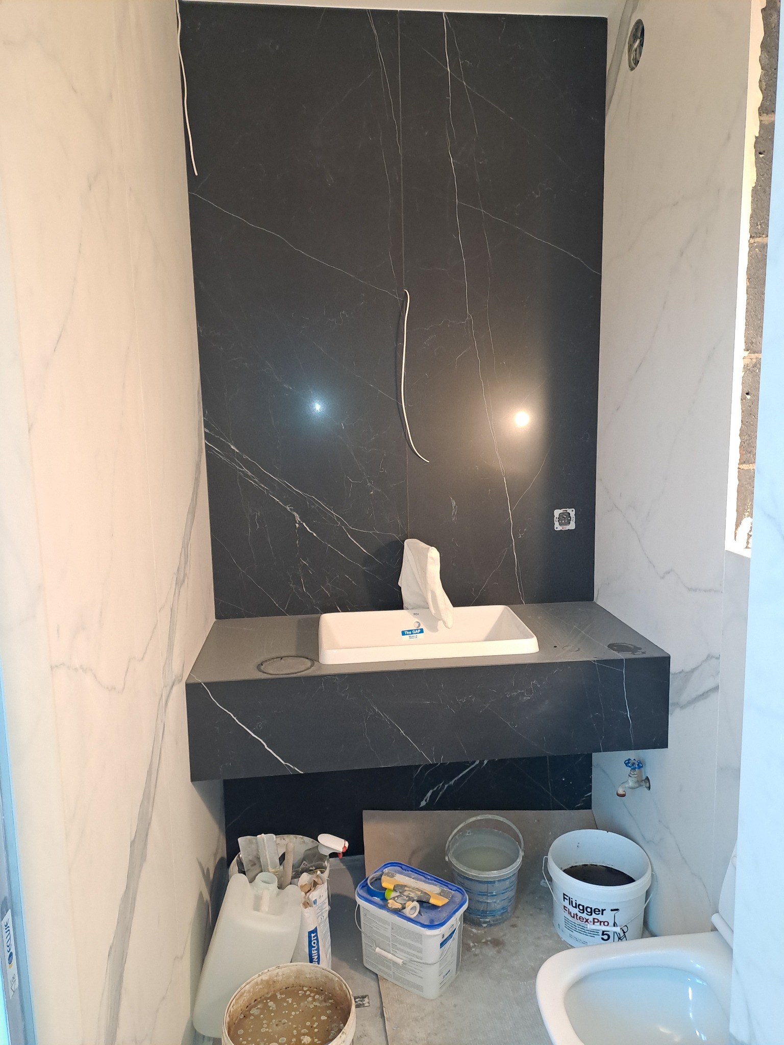 Nowoczesna łazienka w trakcie wykańczania: czarne i białe marmurowe płytki, umywalka w blacie, widoczne narzędzia i materiały budowlane.