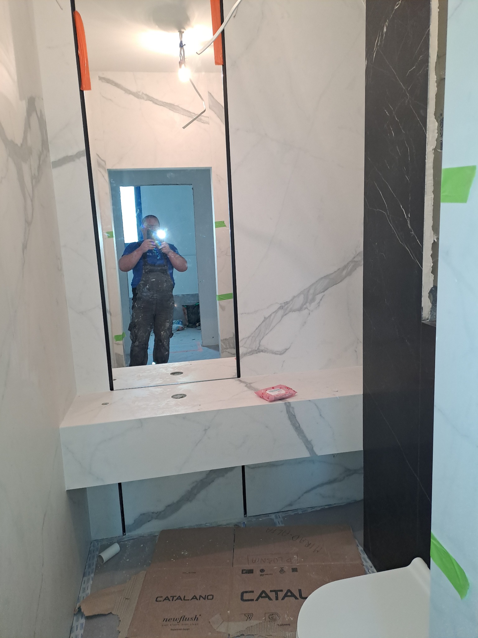 Wykończona łazienka z marmurowymi płytkami, odbicie glazurnika w lustrze. Widoczne kartony po ceramice Catalano. Nowoczesny design z elementami czerni i bieli.