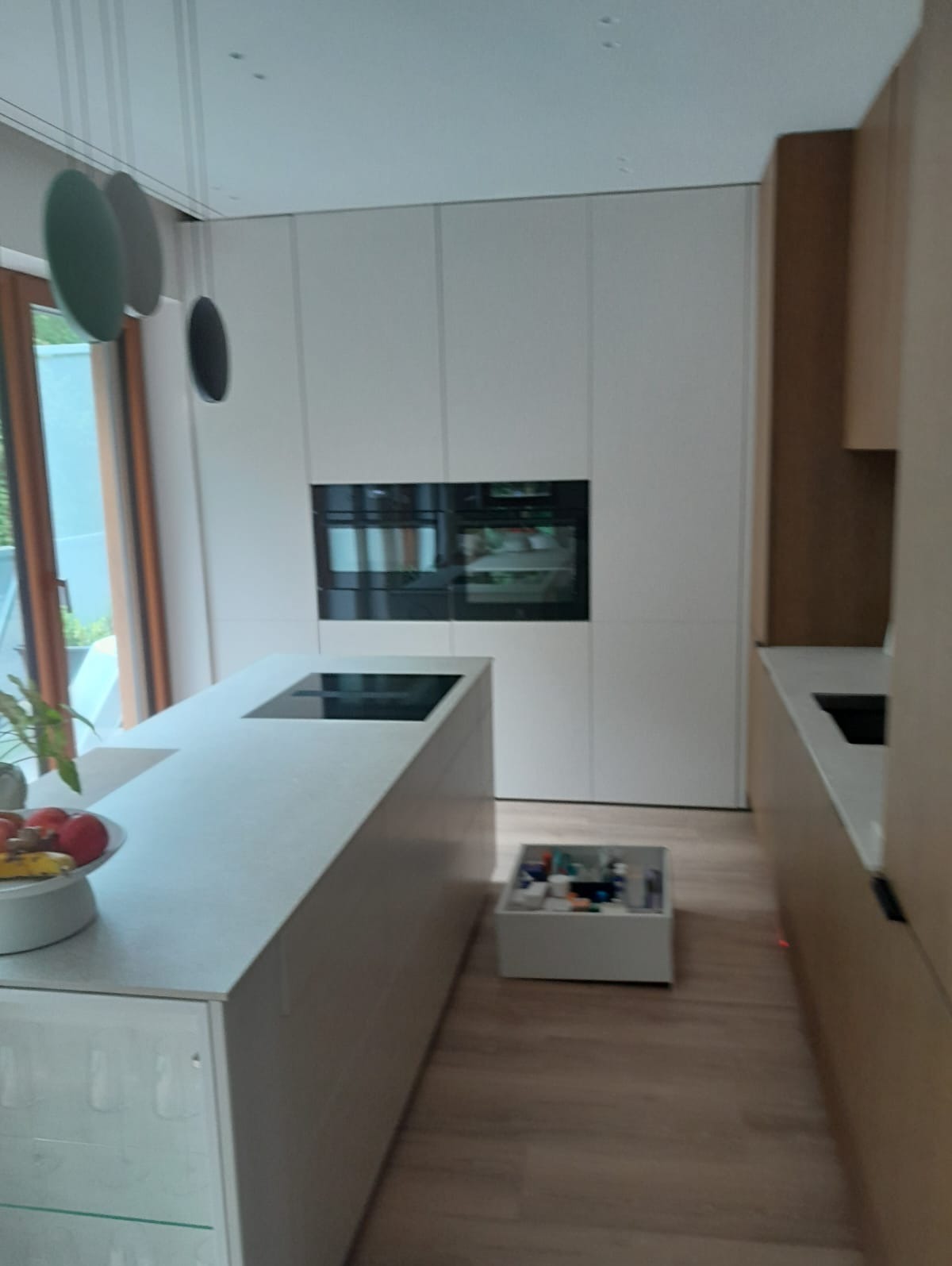 Nowoczesna kuchnia z białymi frontami, wbudowanym piekarnikiem i wyspą z płytą indukcyjną. Minimalistyczny design, drewniana podłoga, oświetlenie punktowe.