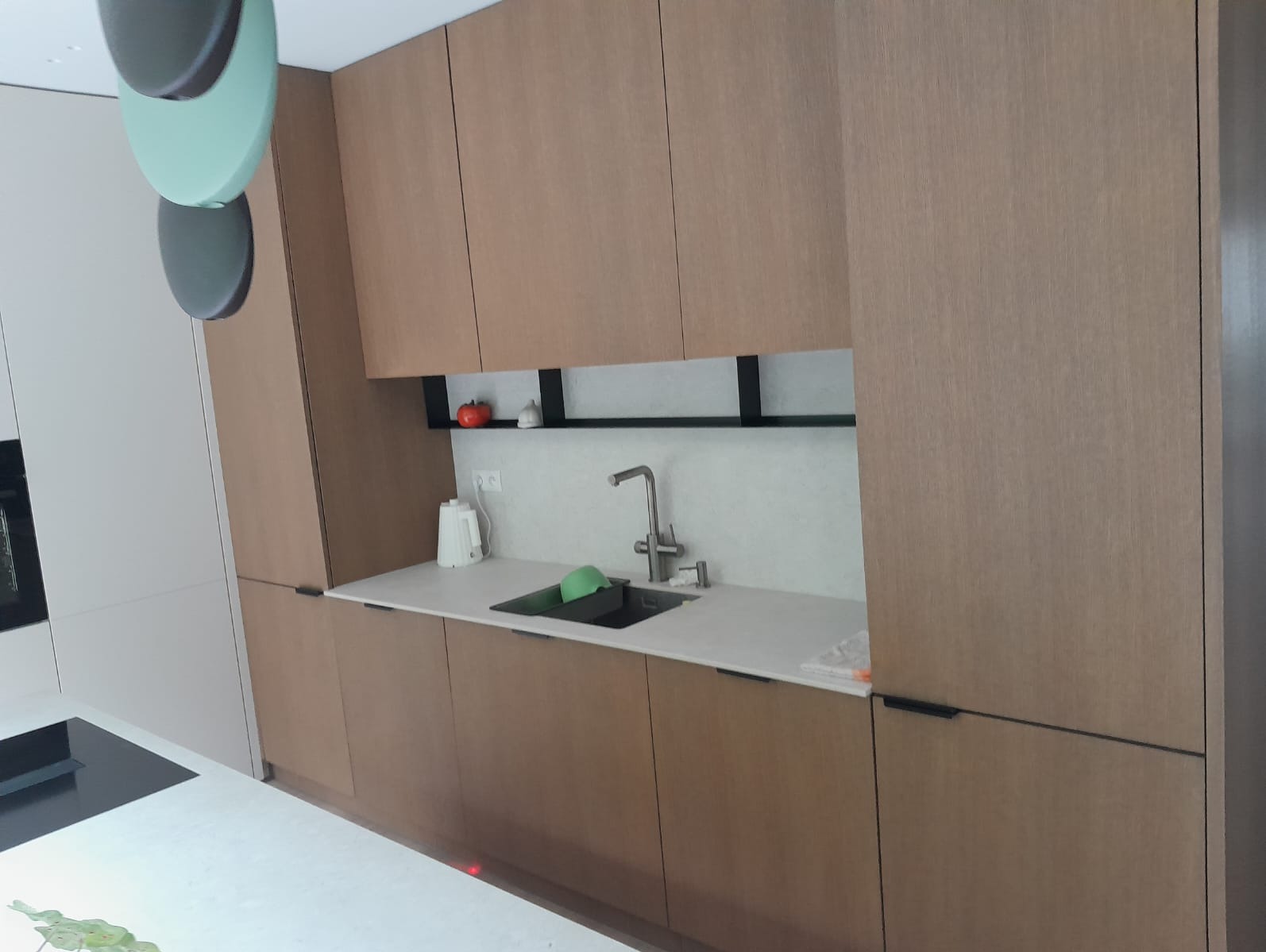 Nowoczesna kuchnia na wymiar z drewnianymi frontami, białym blatem i minimalistyczną armaturą. Widoczny zlew, czajnik i dekoracje na półce. Ujęcie z perspektywy użytkownika.