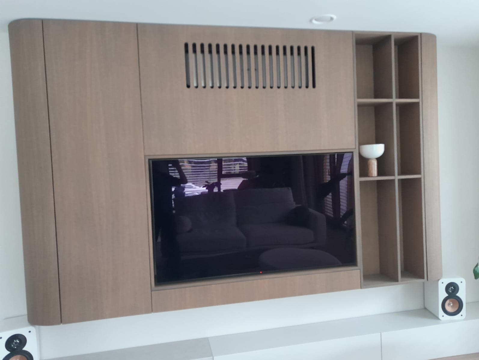 Drewniana zabudowa ściany z wnęką na telewizor i półkami. Widoczne głośniki i odbicie wnętrza w ekranie. Górna część zabudowy z perforacją wentylacyjną.