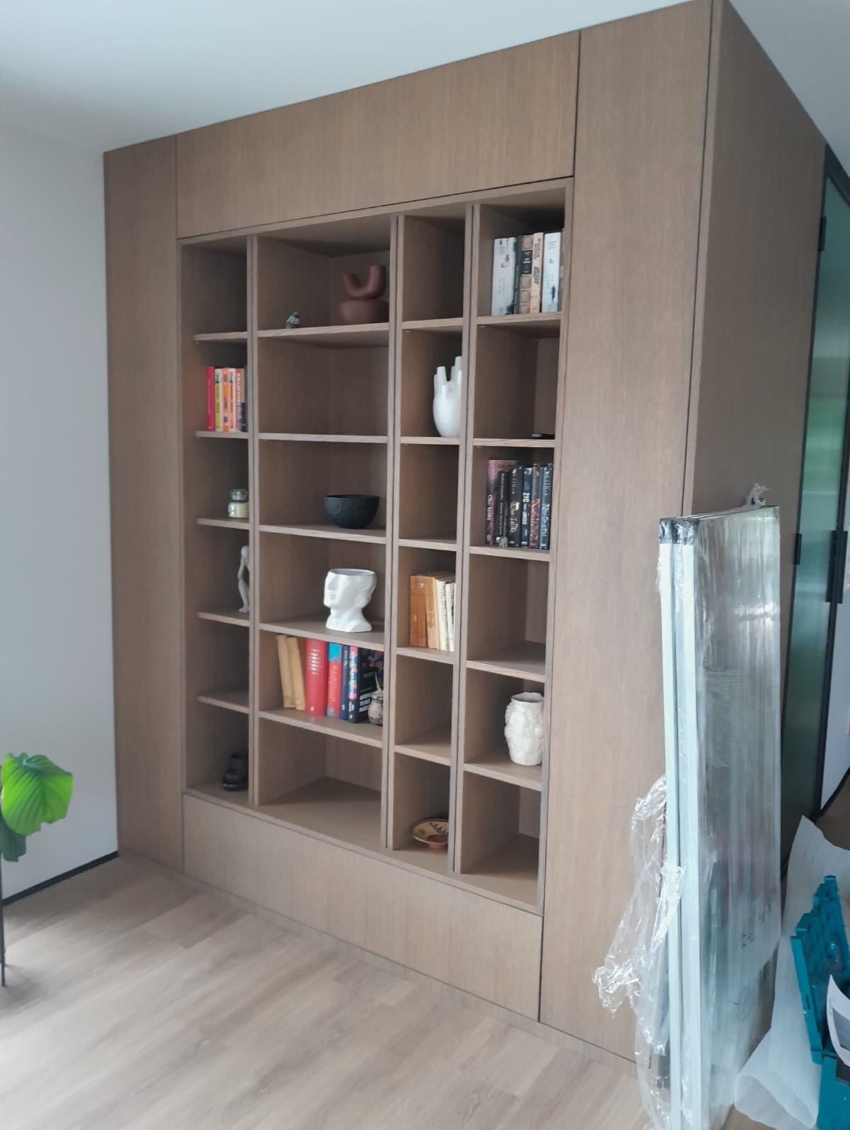 Drewniana szafa wnękowa z półkami, wypełniona książkami i dekoracjami. Widoczne detale wykończenia i oświetlenie wnętrza. Minimalistyczny design.