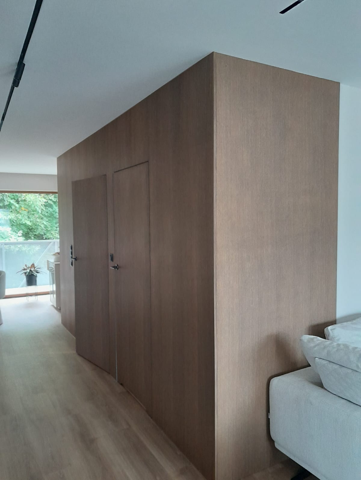 Drewniana zabudowa wnękowa z dwiema parami drzwi, wkomponowana w przestrzeń salonu z widokiem na balkon. Minimalistyczny design, jasna podłoga, naturalne światło.